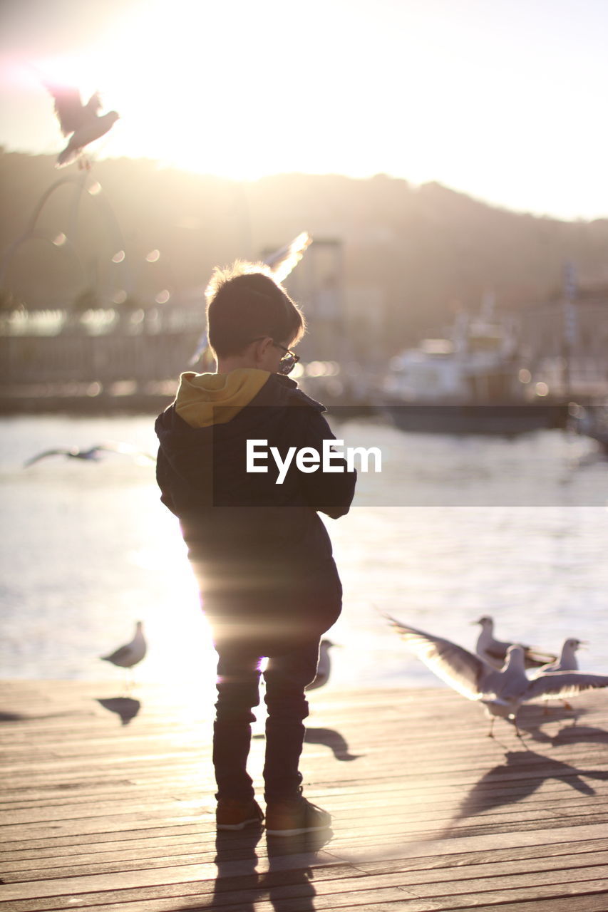 Boy standing on pier over lake against sky during sunset