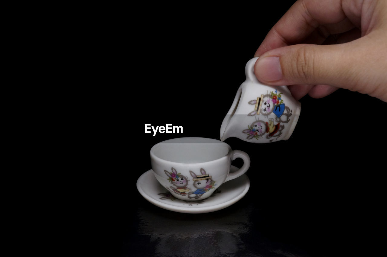 Cropped hand holding jug by empty coffee cup against black background