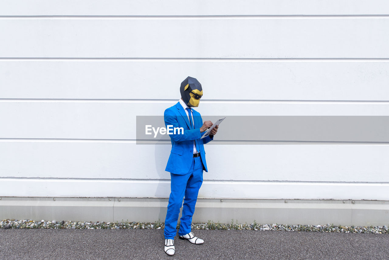 Man wearing vibrant blue suit and monkey mask writing in clipboard in front of white wall