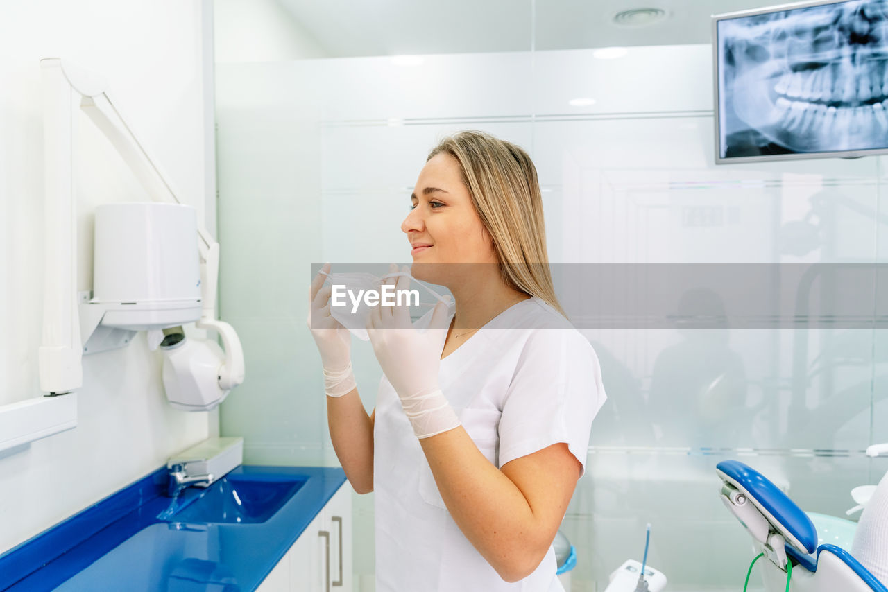 Side view of young female dentist in uniform and gloves putting on medical mask while standing in modern dental clinic