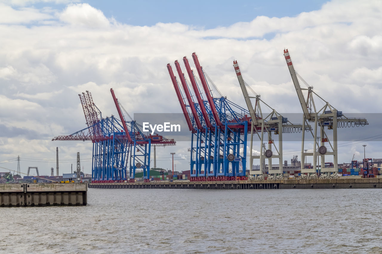 CRANES AT COMMERCIAL DOCK BY SEA AGAINST SKY