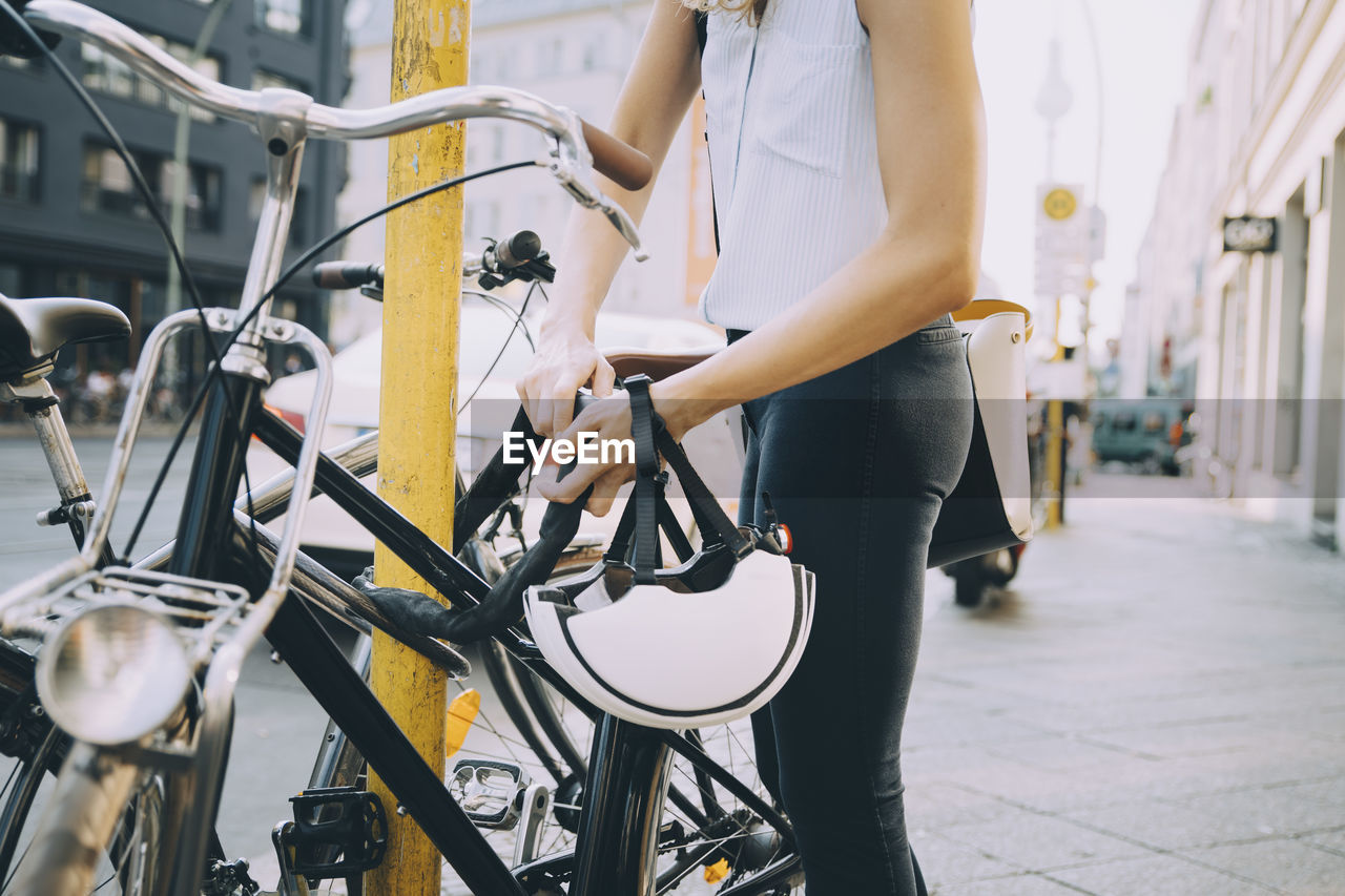 Midsection of businesswoman locking bicycle with pole on sidewalk in city