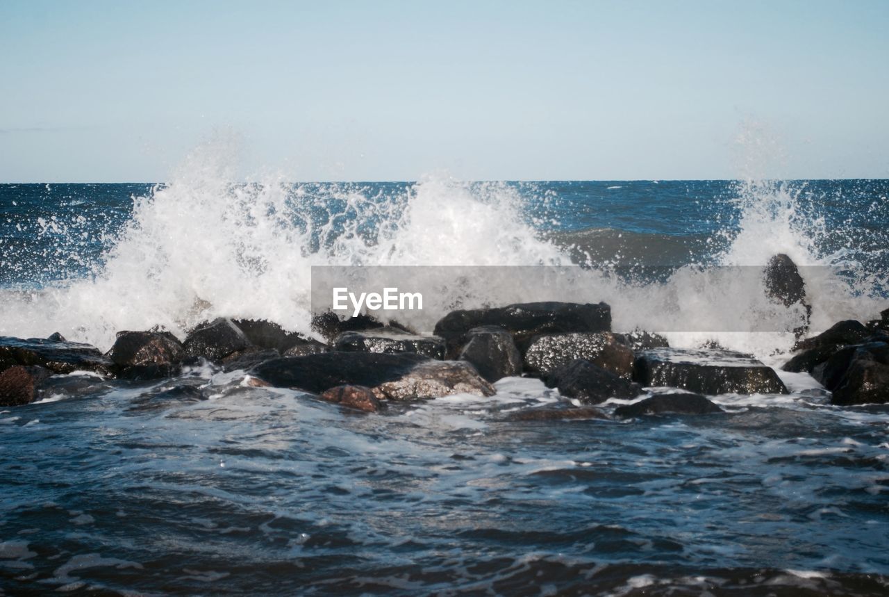 Waves splashing on rocks against clear sky