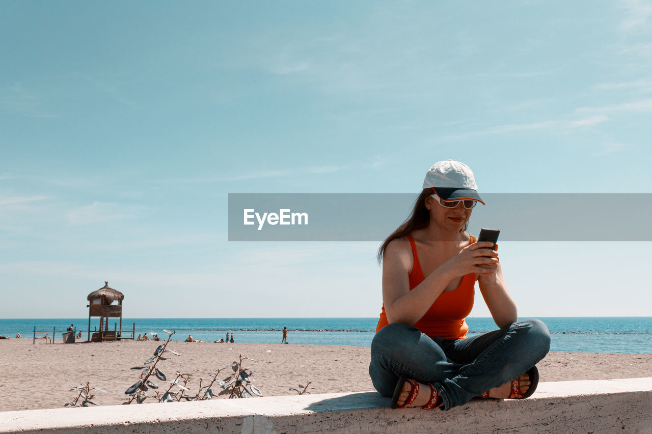 Full length of smiling woman using phone sitting against sea and sky