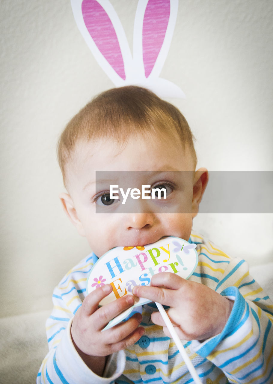 Optical illusion of baby boy with rabbit ears biting toy against wall