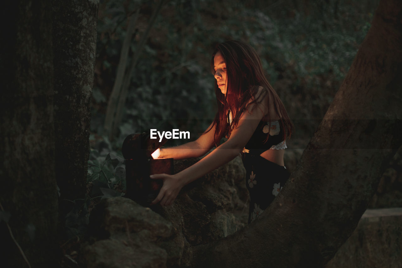 From below of young female in dress taking ancient retro clock in wooden box on stone in autumnal forest