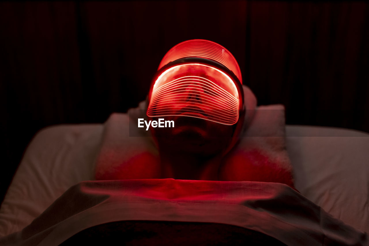 Young woman wearing illuminated red mask during rejuvenating treatment in dark at beauty spa