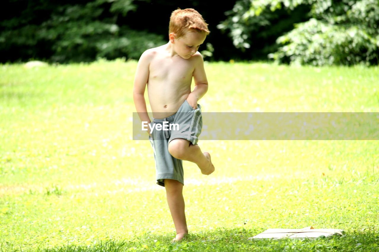 Shirtless boy standing on one leg at park during sunny day