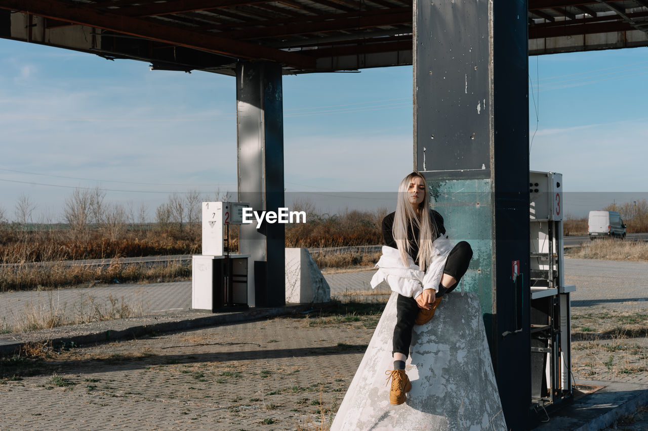 Young woman sitting at fuel pump against sky