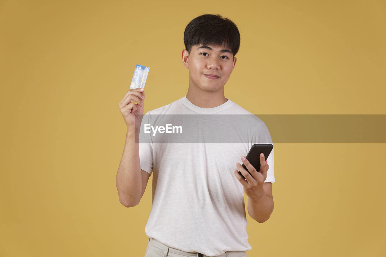 PORTRAIT OF SMILING YOUNG MAN HOLDING CAMERA WHILE STANDING ON YELLOW PHONE