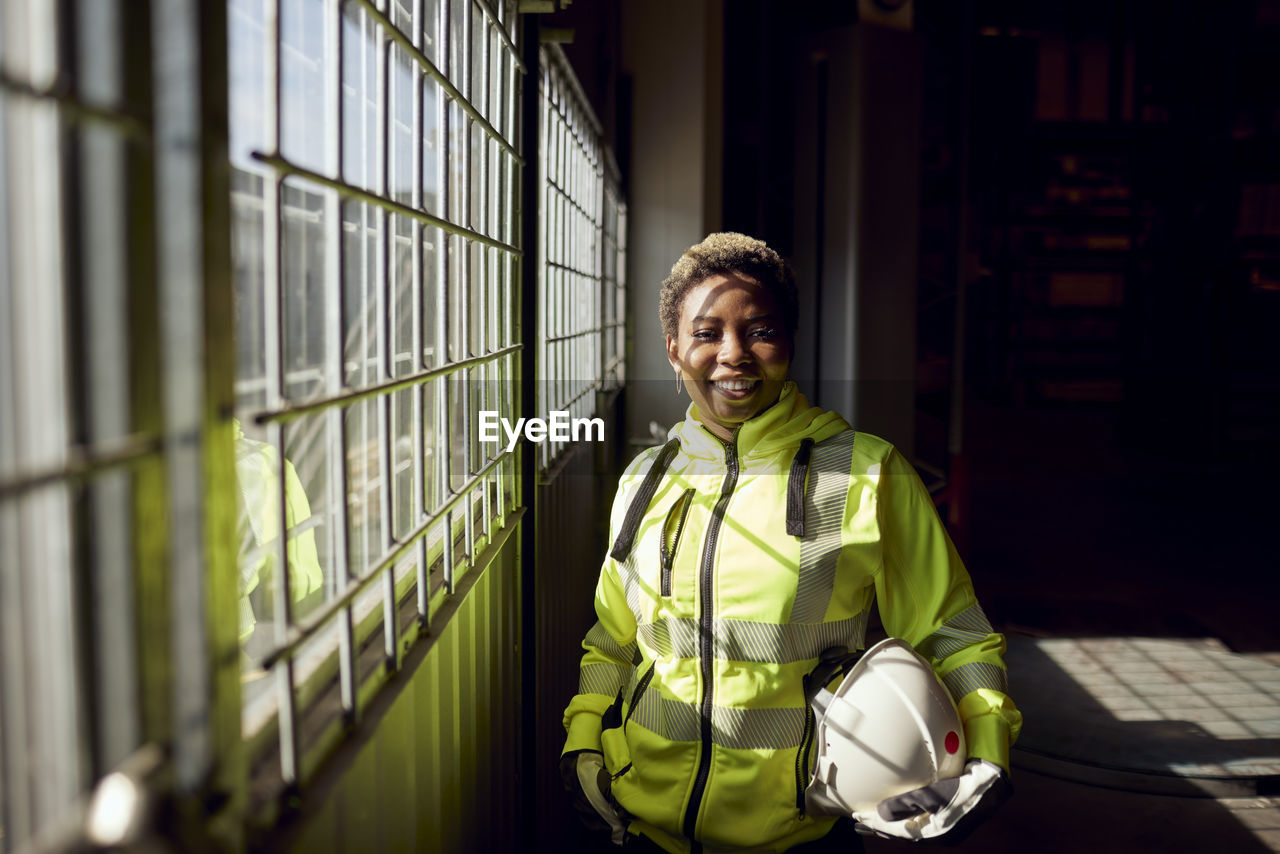 Portrait of smiling young female blue-collar worker standing by window in factory