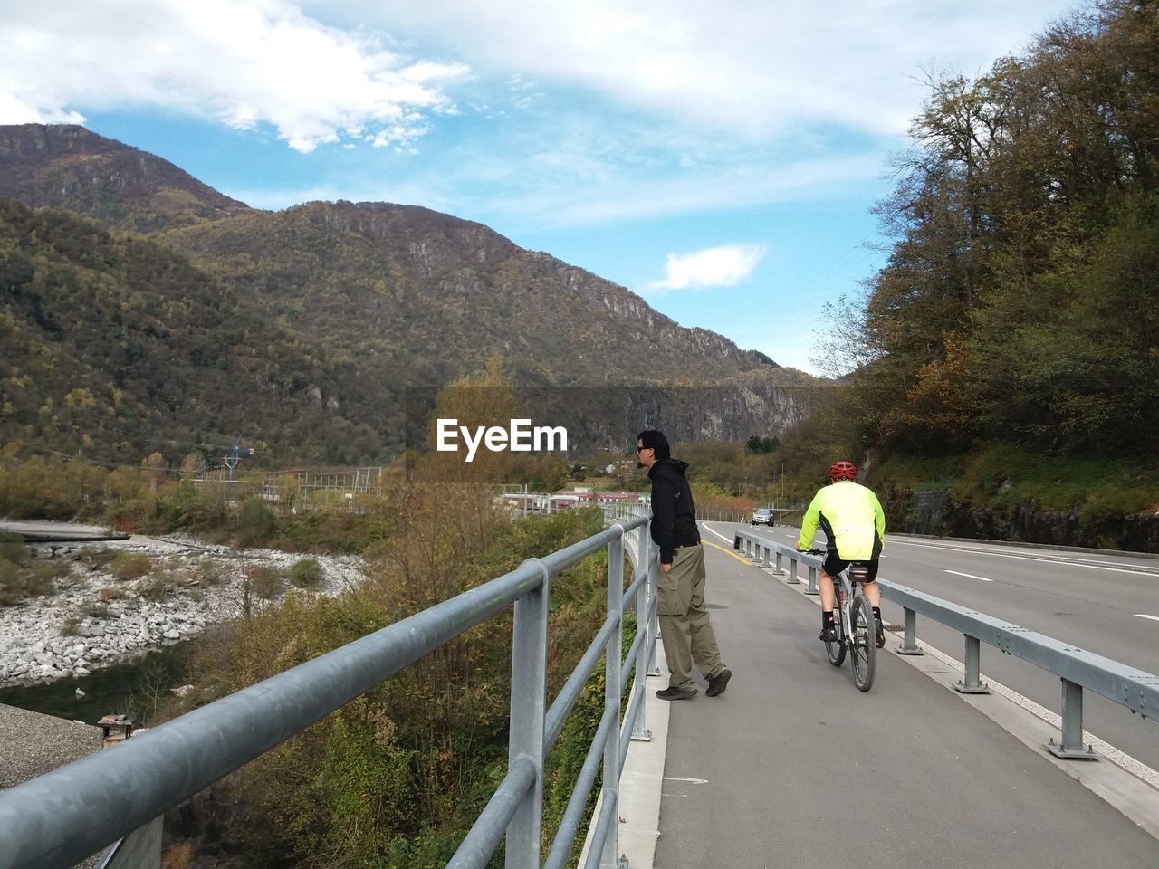 Side view of man standing by cyclist on bridge against mountains