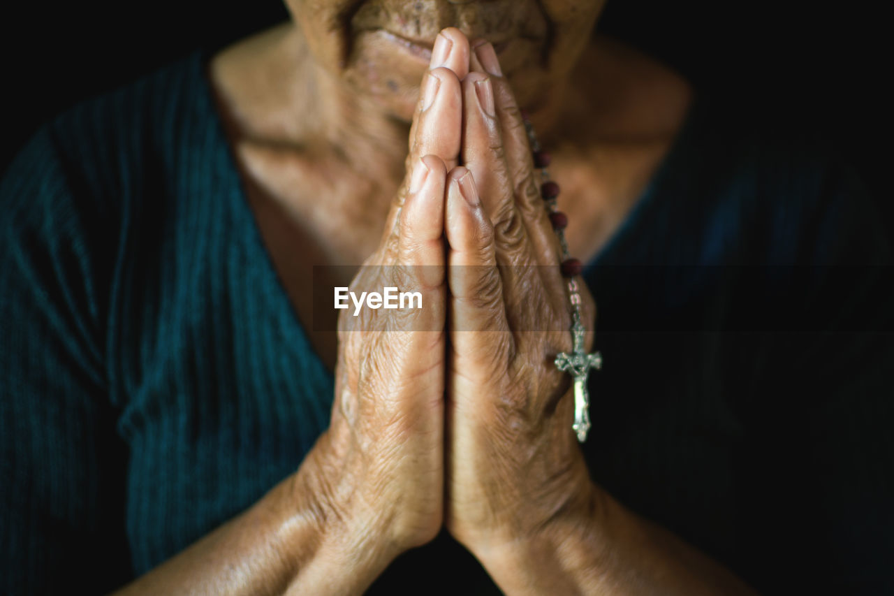 Midsection of senior woman holding rosary praying against black background