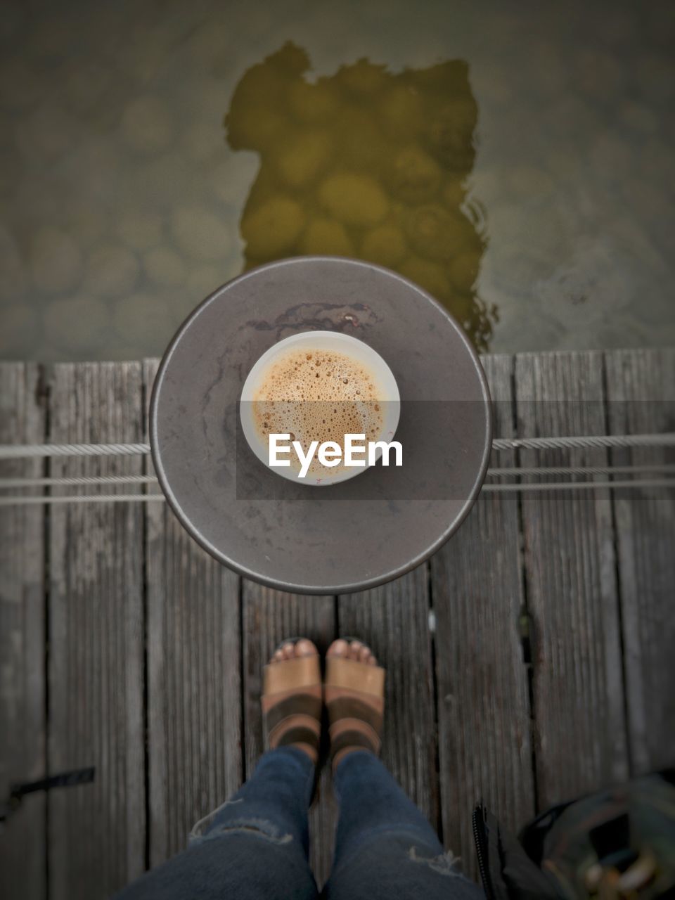 Low section of person standing by coffee cup on pier over lake