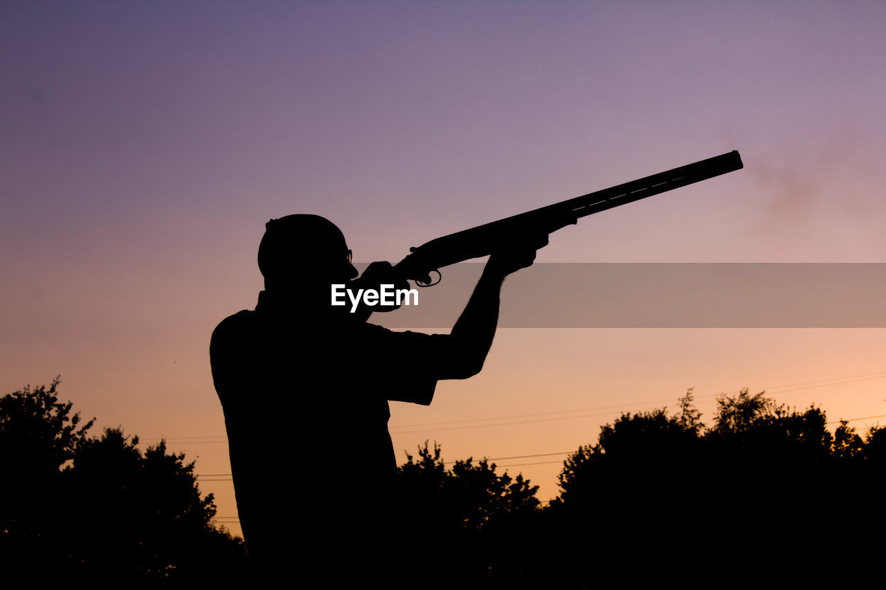 Silhouette man aiming shotgun against clear sky during sunset