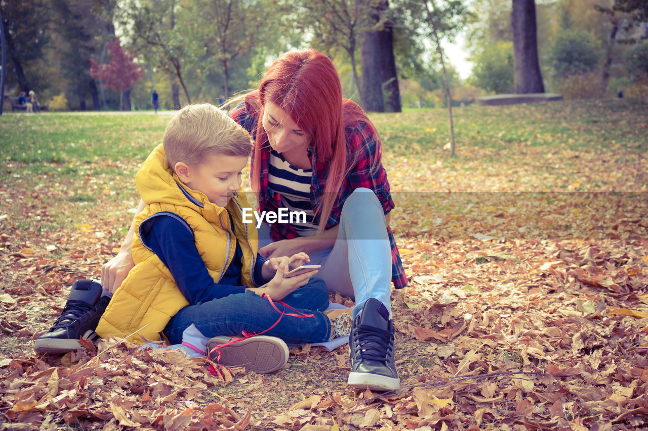 Mother with son using smart phone while sitting on field at park during autumn