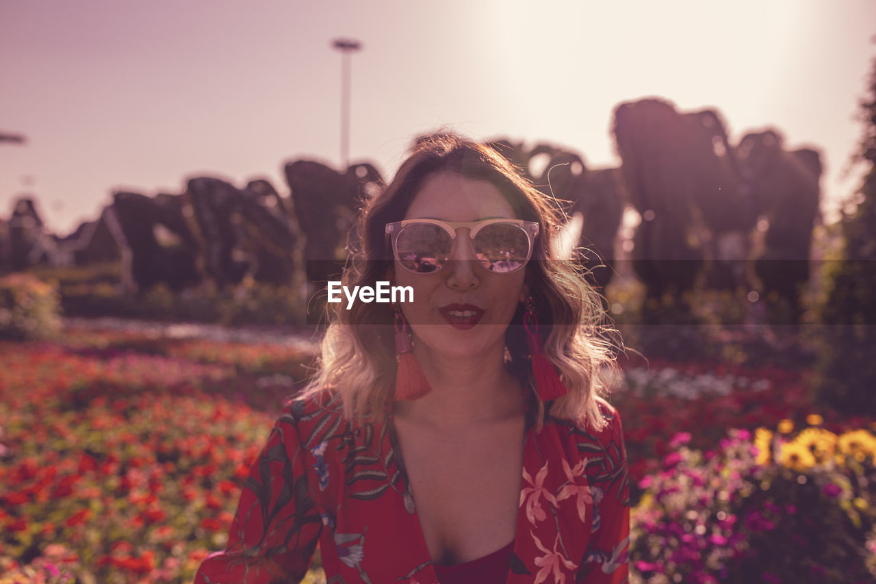 Portrait young woman standing in ornamental garden against sky at dusk