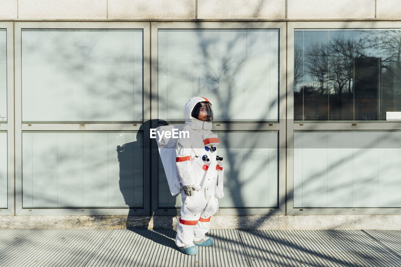 Young female astronaut in space suit standing by glass window in city during sunny day