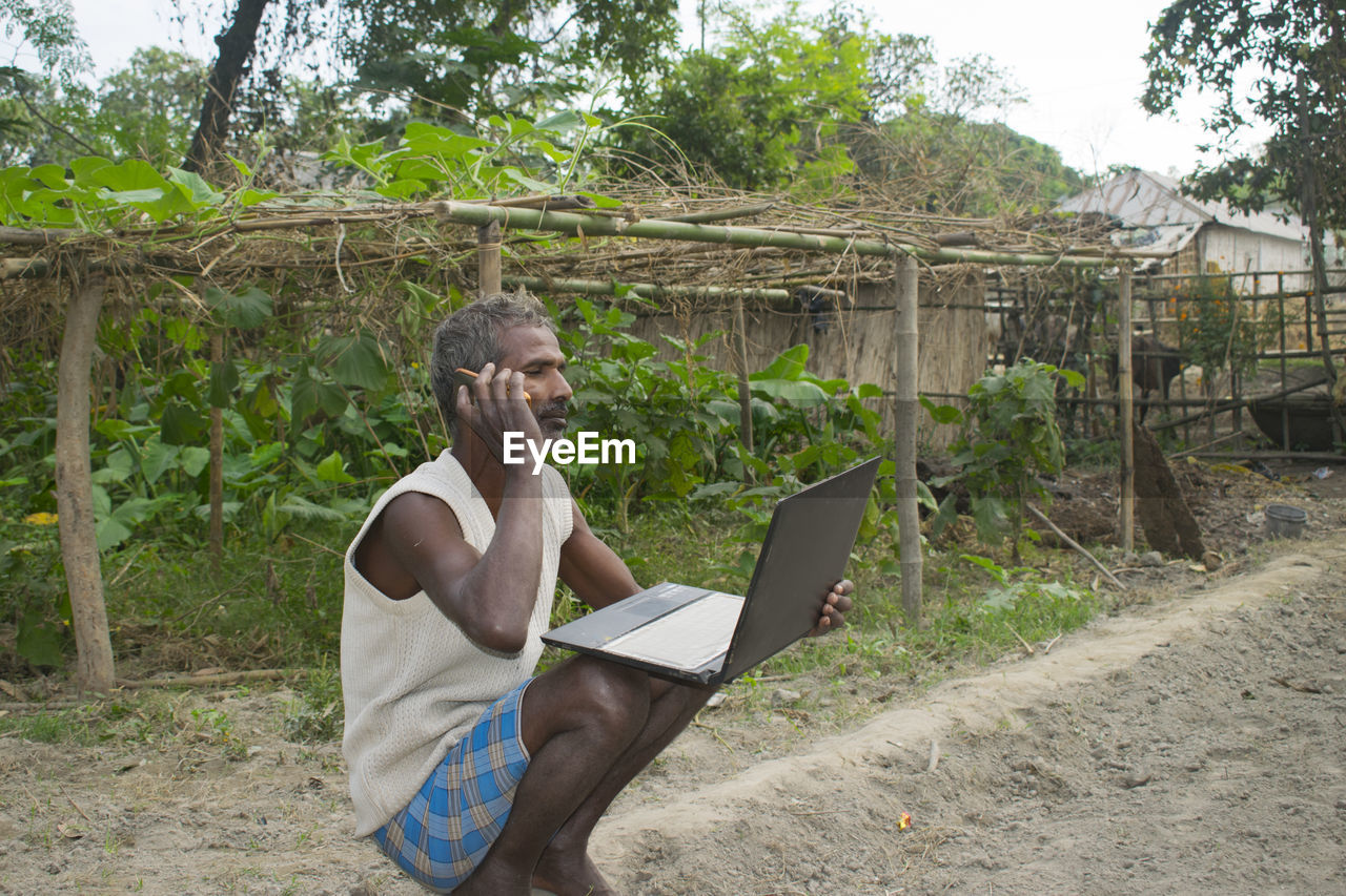 REAR VIEW OF MAN USING MOBILE PHONE IN GARDEN