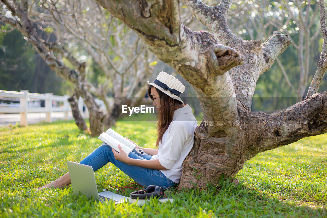 Side view of woman reading while sitting by tree trunk at park