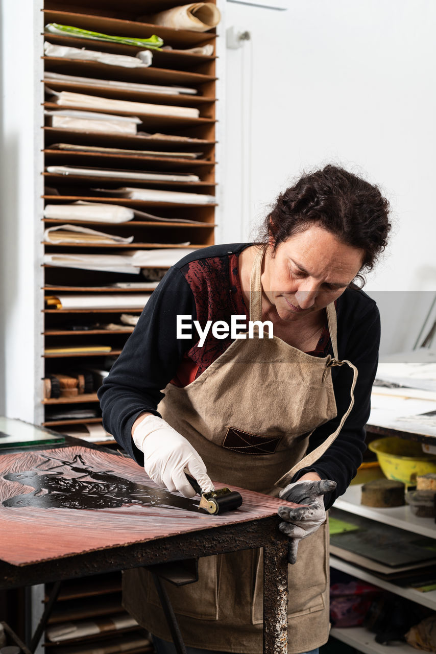 Mature female artist in apron and gloves covering engraved plate with ink while creating artwork in professional workroom