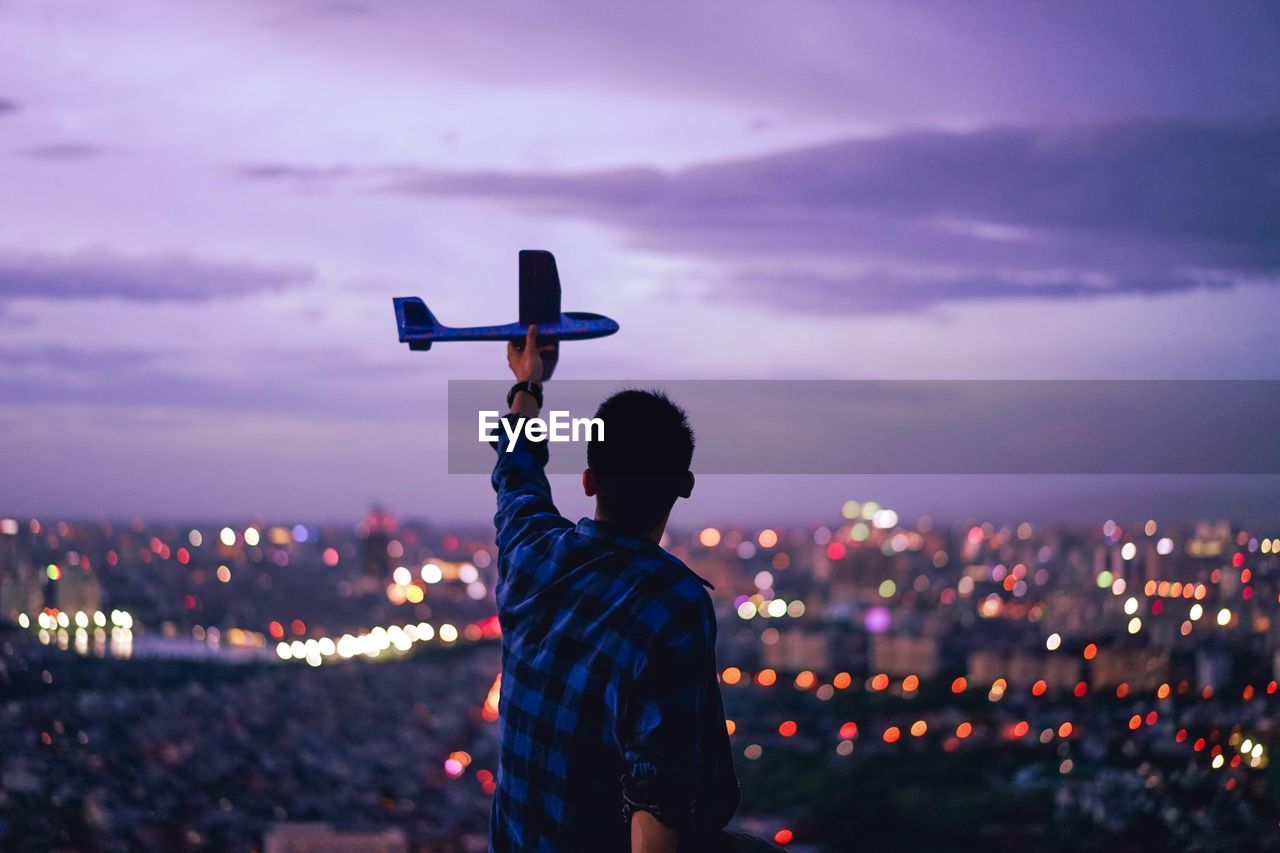 Rear view of man holding model airplane against illuminated cityscape at dusk