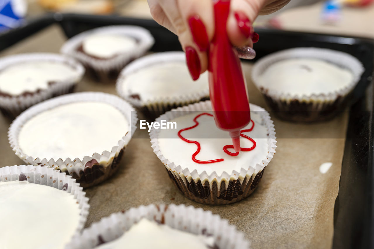 A woman squeezes colored frosting from a tube onto chocolate brown cupcakes covered  white frosting.
