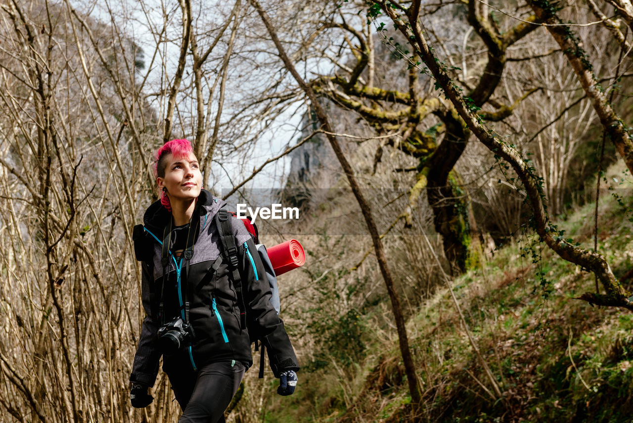Female backpacker with photo camera and backpack trekking among trees and admiring nature