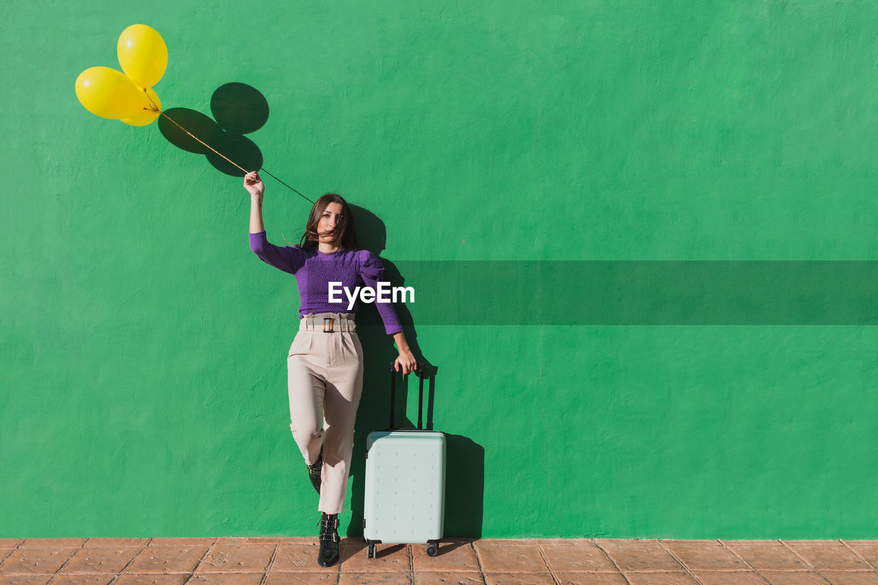 Modern fashionable carefree female holding bunch of yellow balloons and suitcase while standing against green wall in sunny day