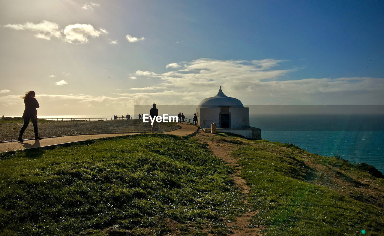 Tourists walking on mountain by chapel at shore against sky in cabo espichel