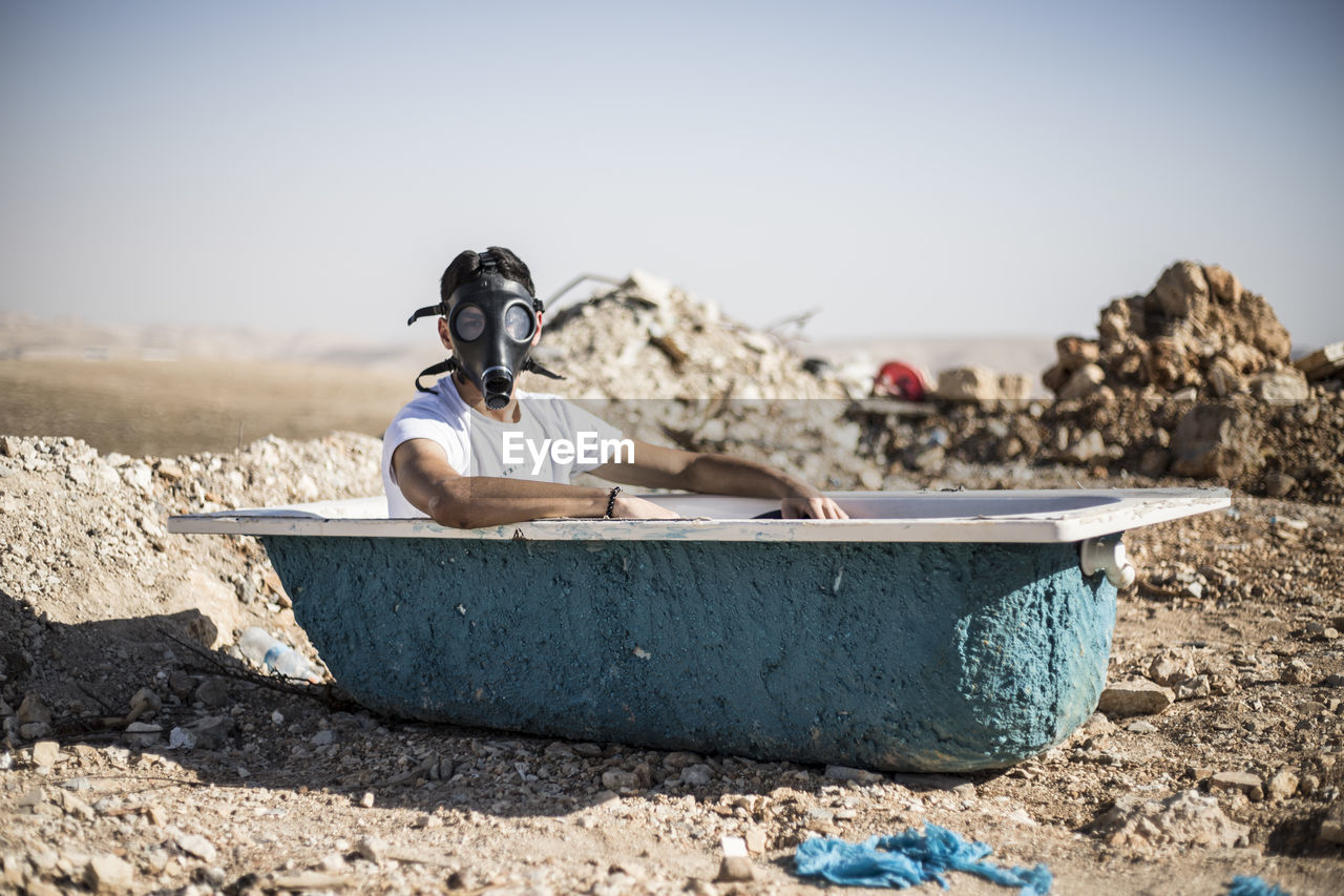 Portrait of young man wearing gas mask in bathtub against clear sky