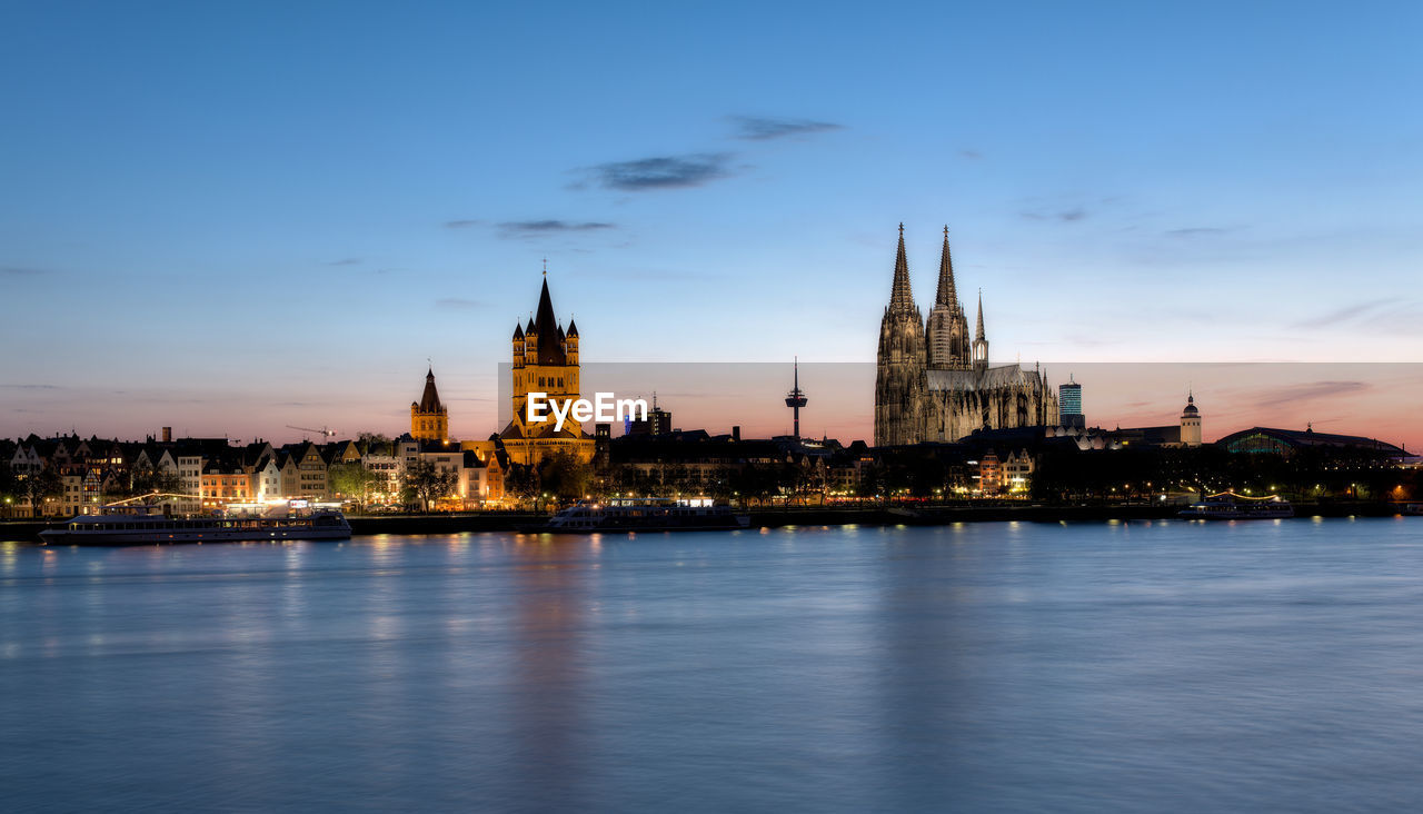 Illuminated buildings and cologne cathedral by rhine river in city at dusk