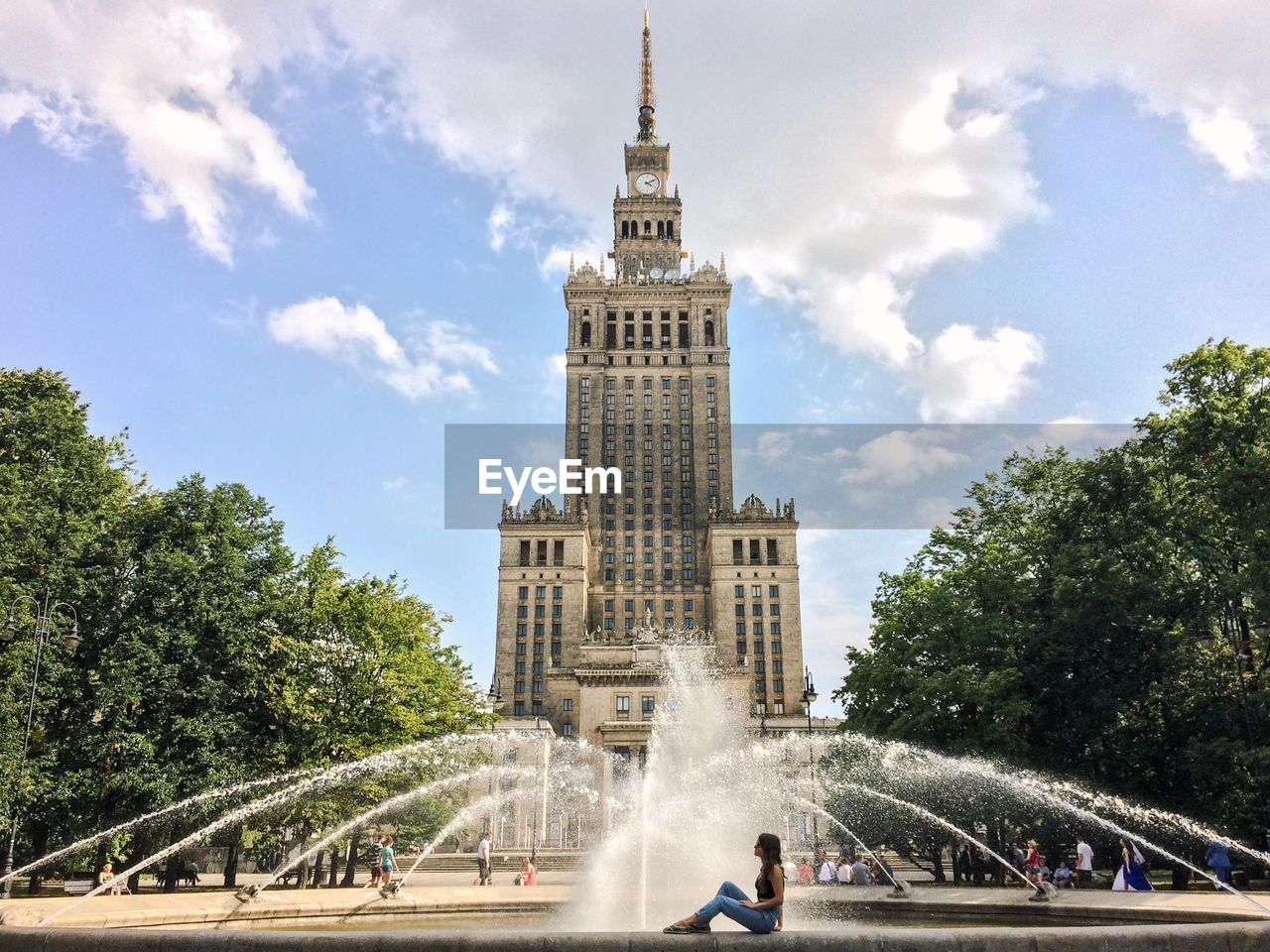 Side view of woman sitting by fountain in front of palace of culture and science