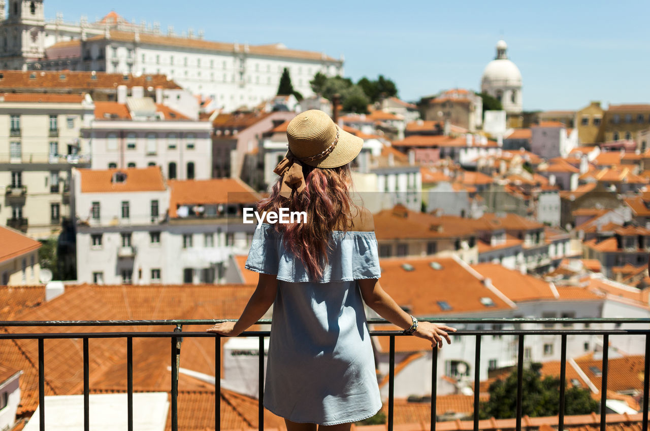 Rear view of young woman looking at buildings while standing by railing in balcony against clear sky