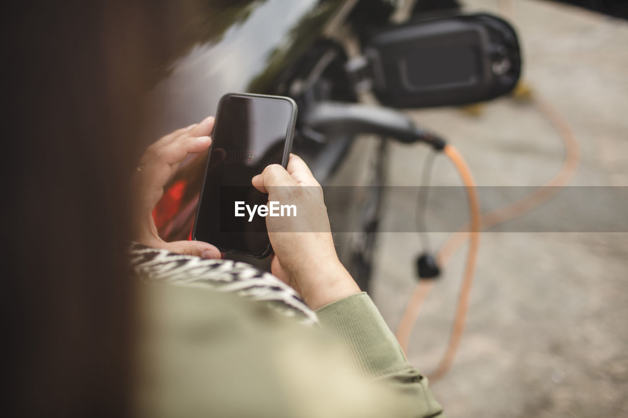 Midsection of woman using smart phone while standing by charging electric car