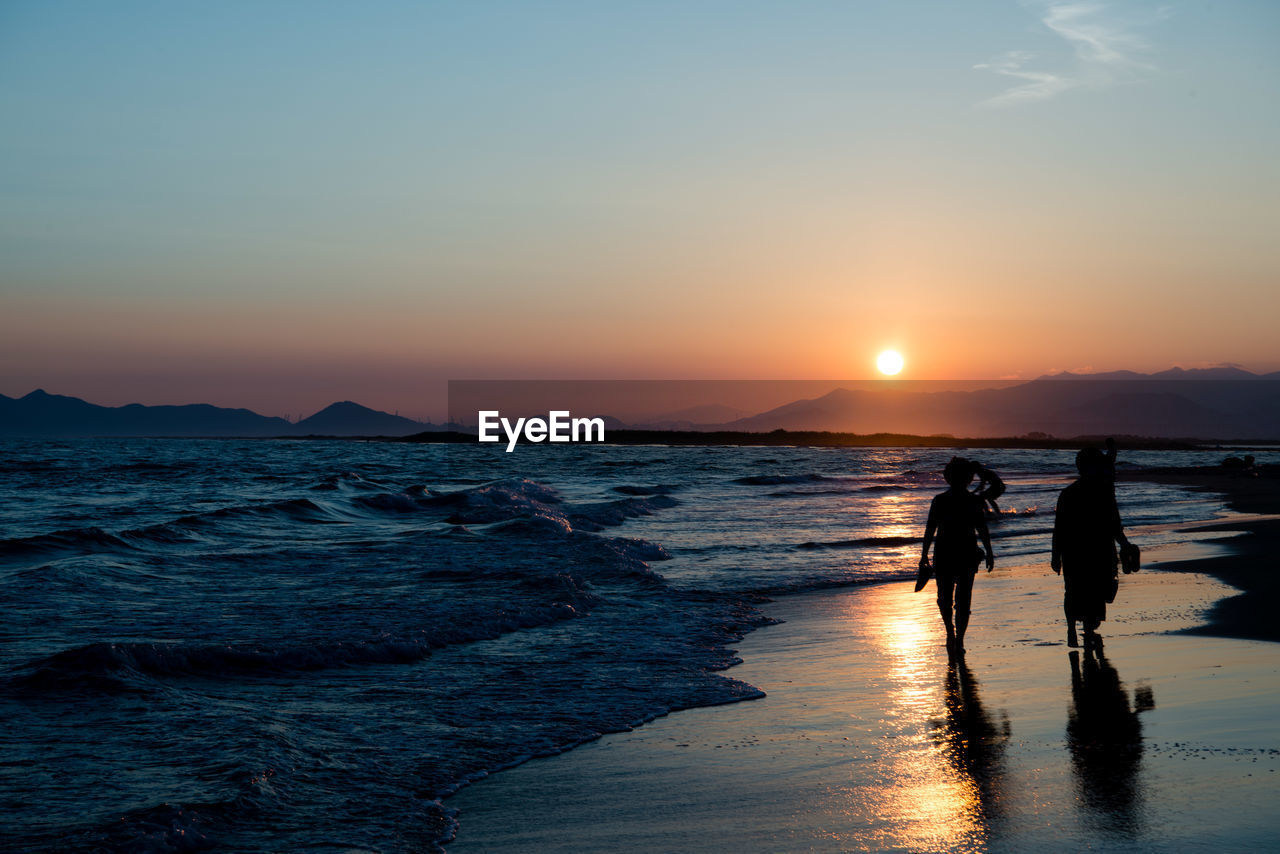 Silhouette people walking on shore at beach against sky during sunset