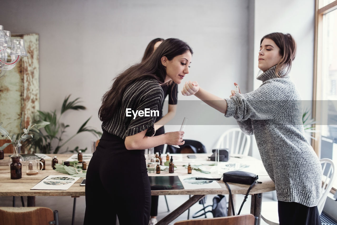 Young woman smelling perfume fragrance from female colleague's wrist while standing at workshop