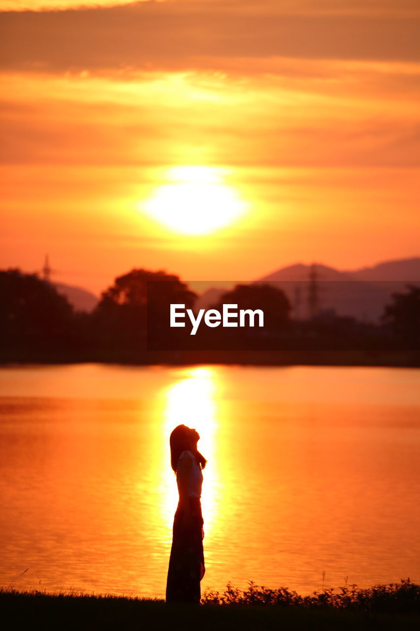 Silhouette woman standing at lakeshore against orange sky