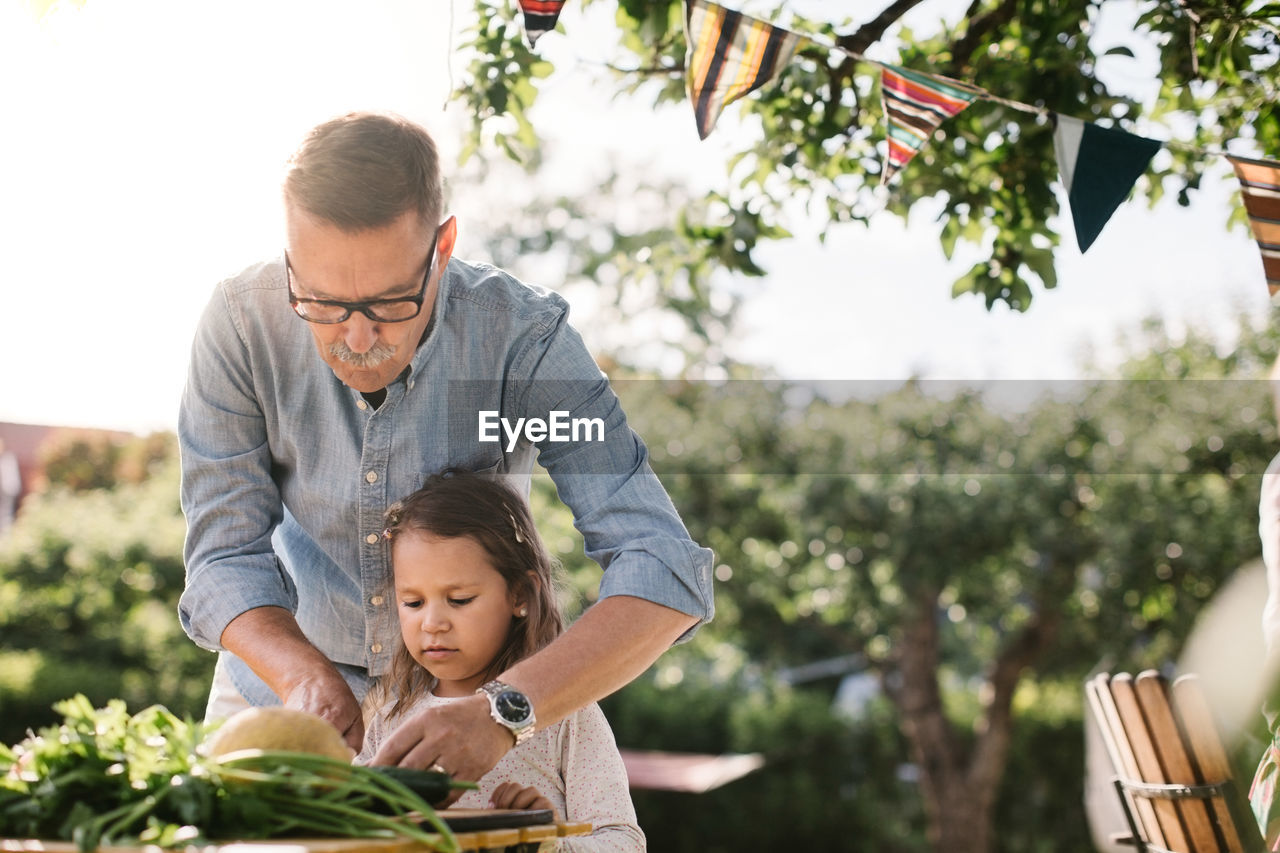 Grandfather teaching granddaughter to cut vegetable at table in backyard