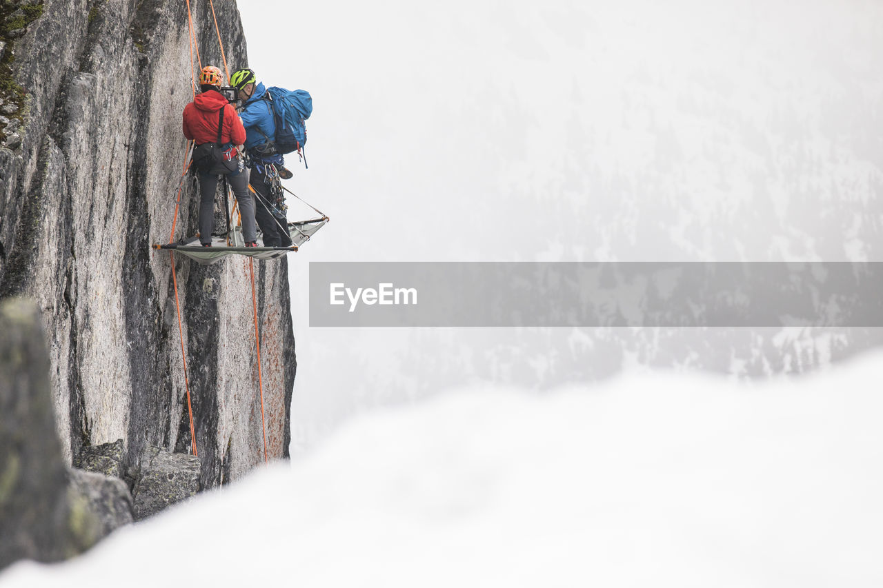 Two climbers standing on portaledge, setting up camp for night.