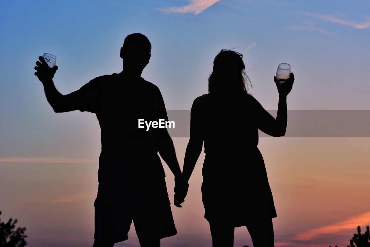 Silhouette couple with wineglasses standing against sky during sunset