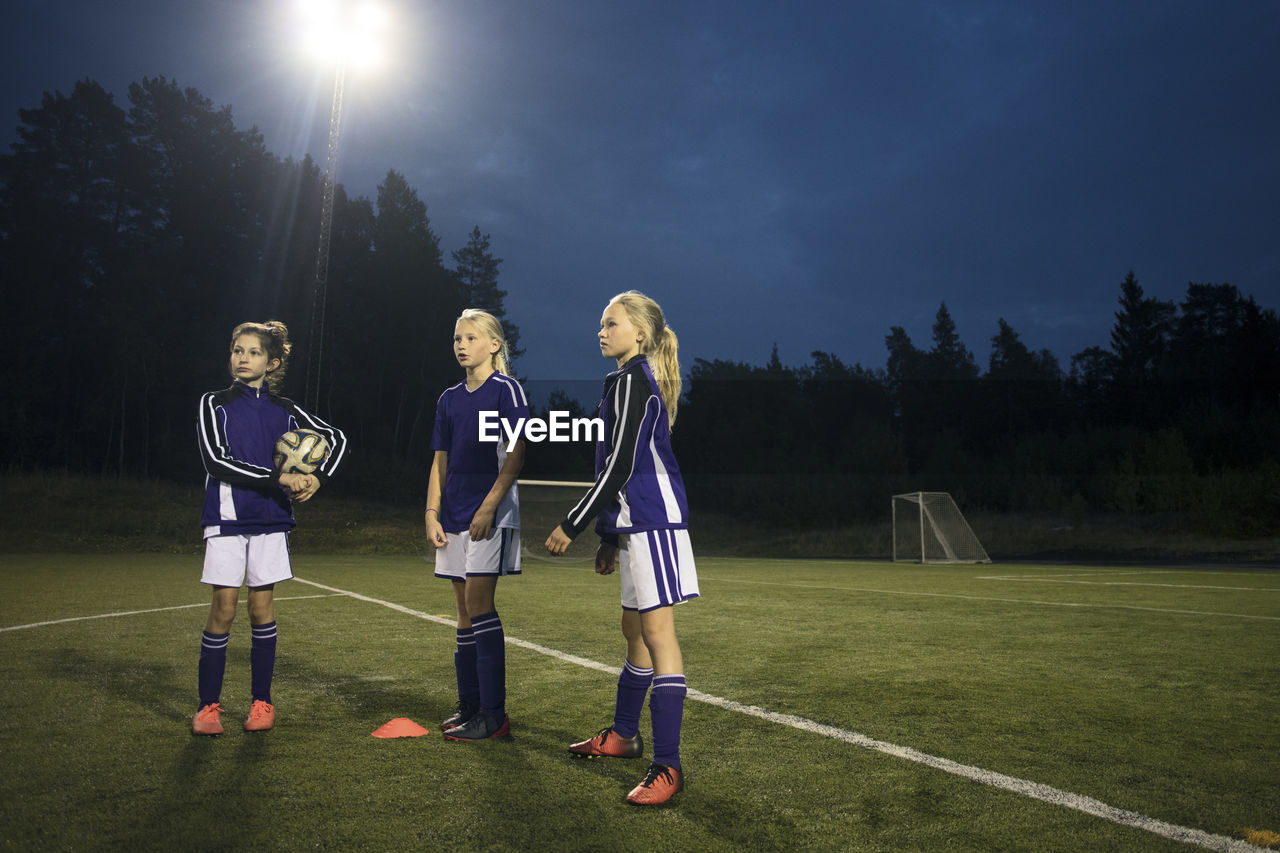 Girls standing on soccer field against sky at dusk