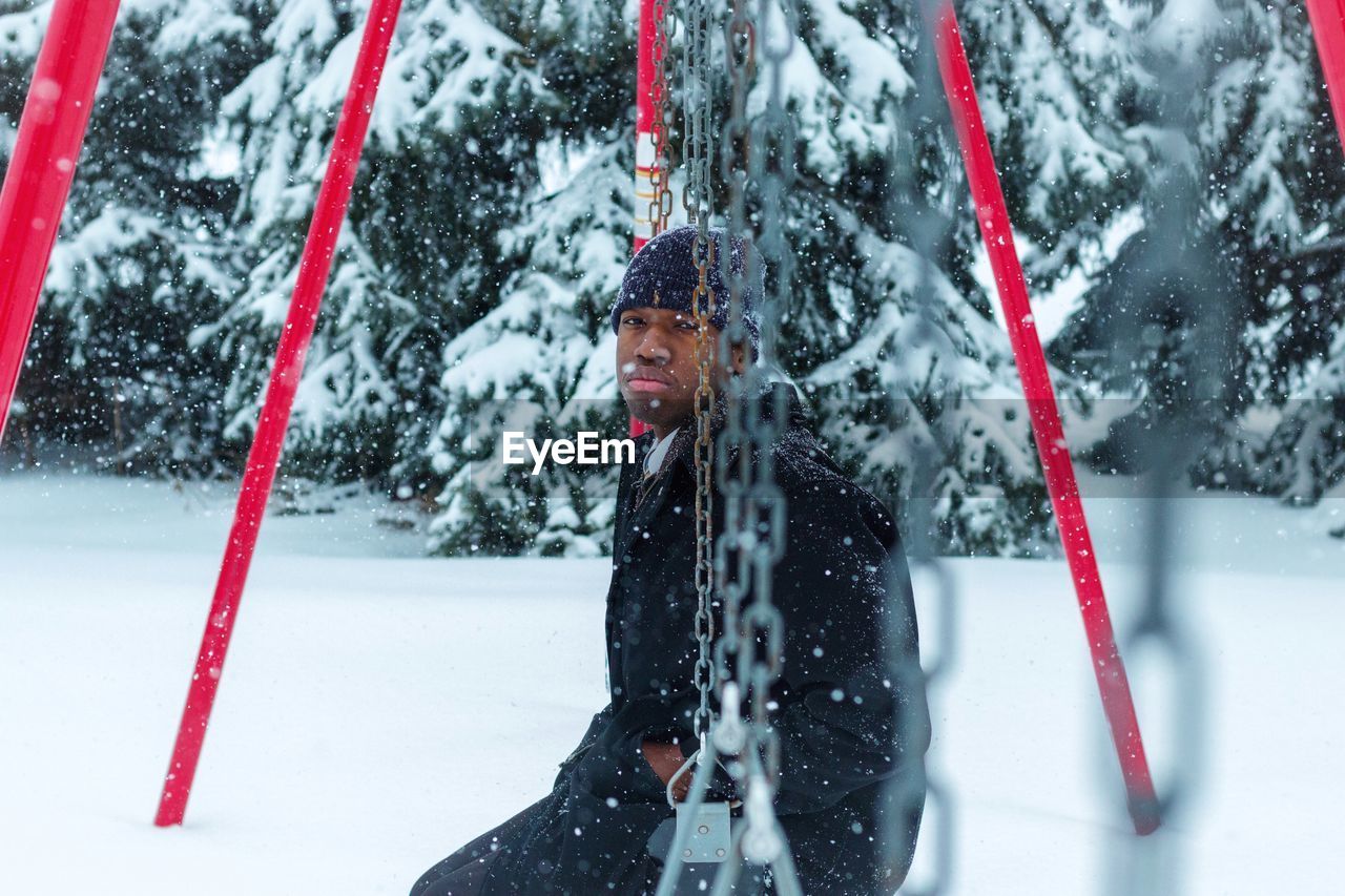 Portrait of man sitting on swing during snowfall
