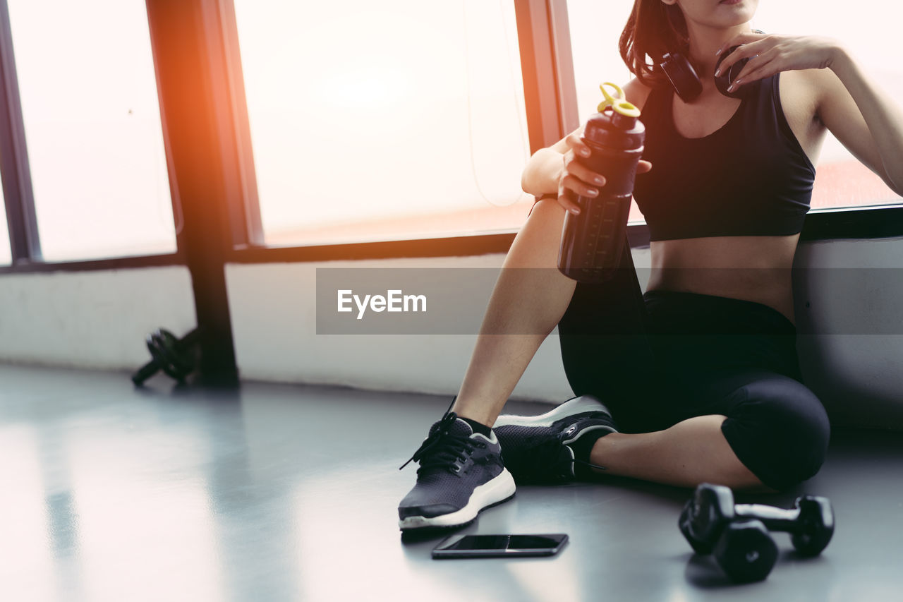 Low section of young woman having protein drink against window in gym