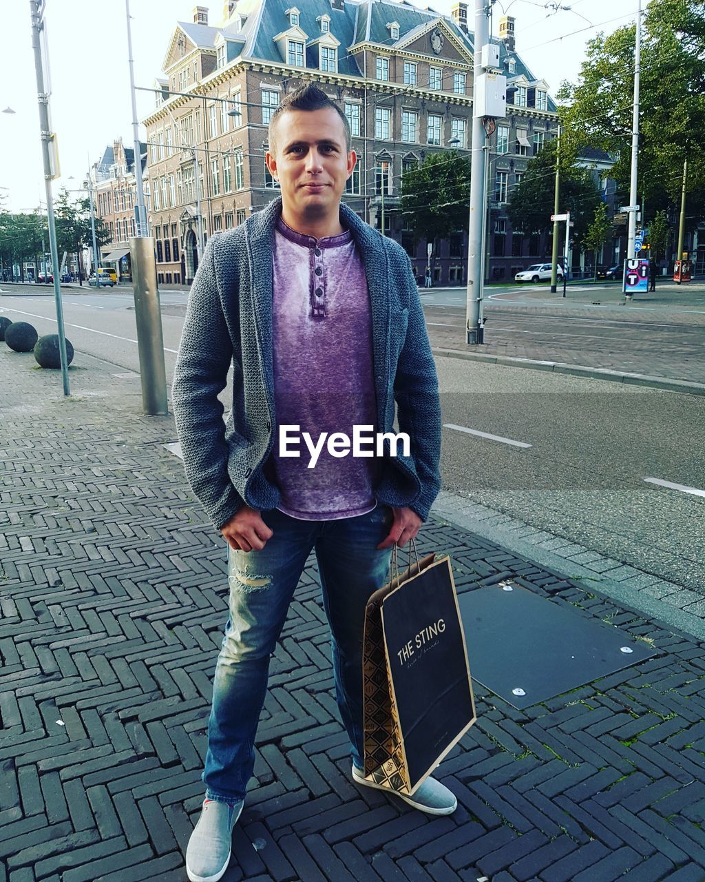 FULL LENGTH PORTRAIT OF YOUNG MAN STANDING ON SIDEWALK IN CITY