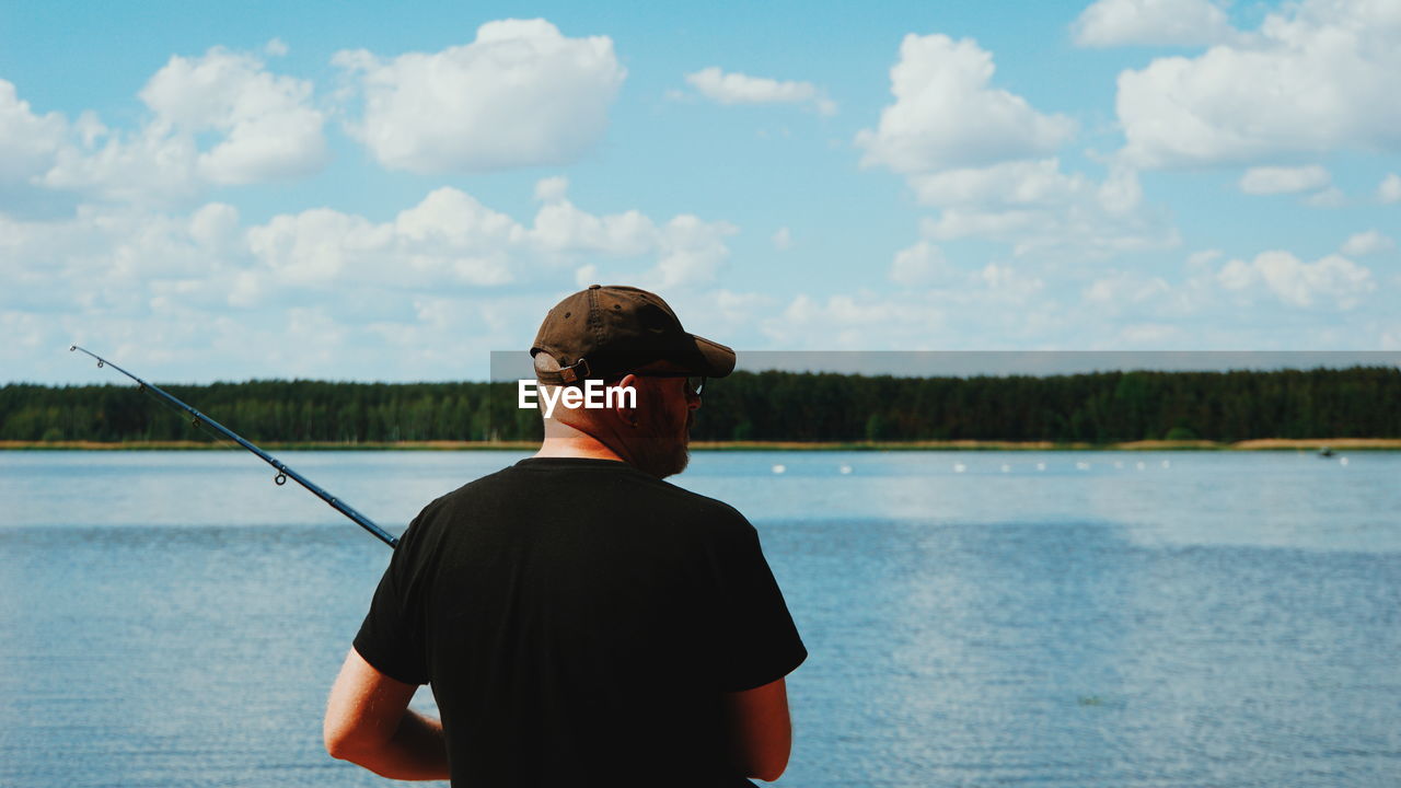 Rear view of man holding fishing rod while standing by lake