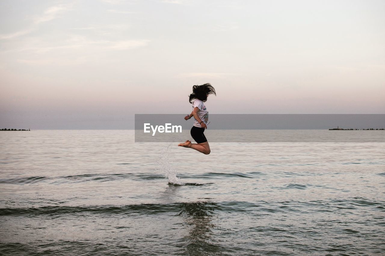 Woman jumping in sea against sky during sunset