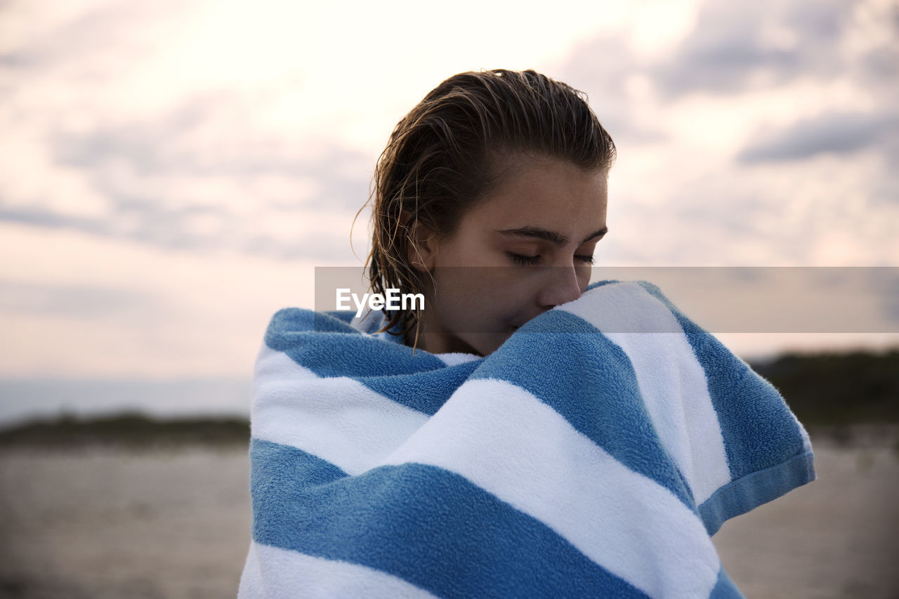 Woman with eyes closed wrapped in towel standing against sky