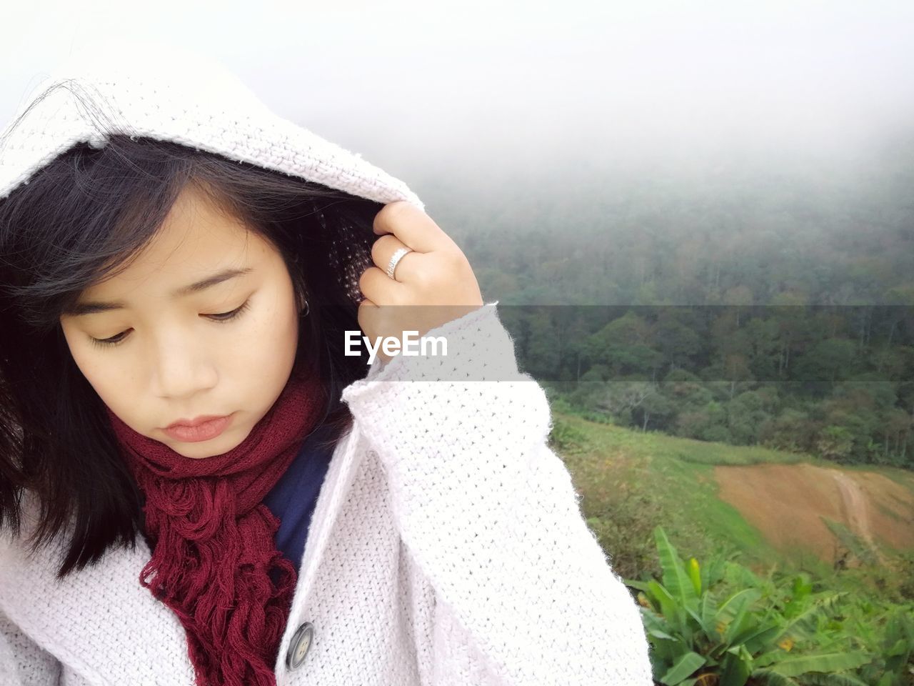 Thoughtful young woman wearing hooded sweater against green landscape