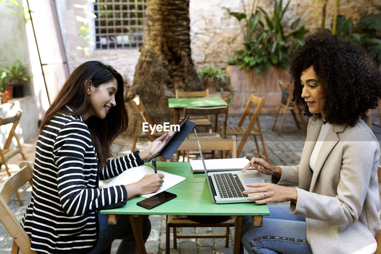 Businesswoman writing in book while female coworker using laptop sitting at cafe